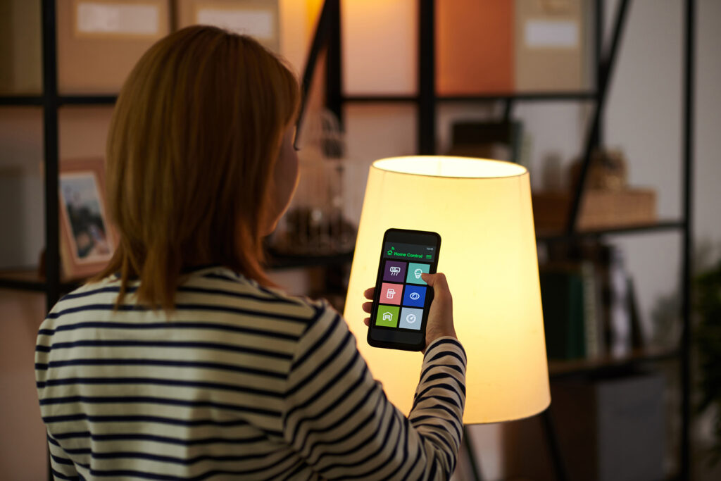 Frau, die vor ihrer Stehlampe steht und diese über ihr Smartphone mit dem Smart Home System bedient.